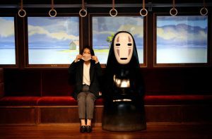 girl sitting besides a life size model of no face from spirited away