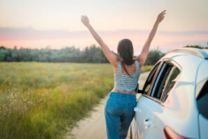 A woman stands beside a car on a summer field road, arms raised in freedom, enjoying a beautiful sunset.