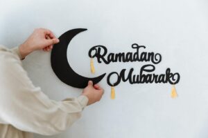 Close-up of hands hanging a Ramadan Mubarak sign with a crescent moon on a wall, signifying festive decoration.